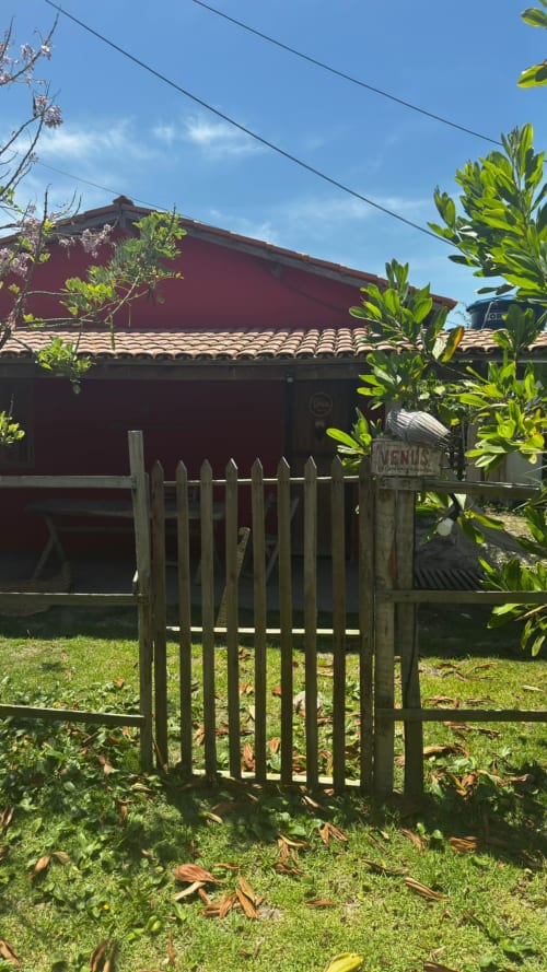 Portão de madeira na entrada da Caraiva Cadu House à beira-mar em Caraiva, Brasil, com chalé vermelho visível entre a vegetação exuberante