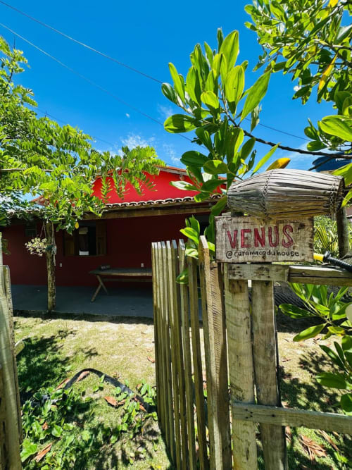 Casa Vênus em Caraiva Cadu com apartamento frente-mar de telhado vermelho, portão de entrada em madeira, plantas tropicais exuberantes e céu azul cristalino