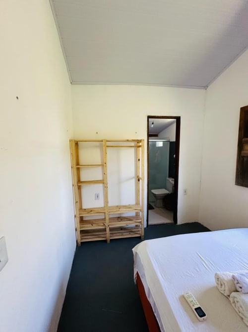 Quarto com guarda-roupa de madeira, cama com roupas brancas, piso escuro e entrada do banheiro na Caraiva Cadu House