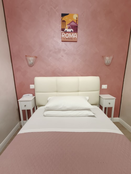 Modernes Schlafzimmer des Hotel Principe Amedeo Rom mit rosa Wänden, cremefarbenem Bett, weißen Nachttischen und Rom-Reiseplakat