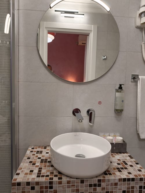 Bagno moderno dell'Hotel Principe Amedeo con specchio circolare, lavabo bianco e pavimento in piastrelle mosaico