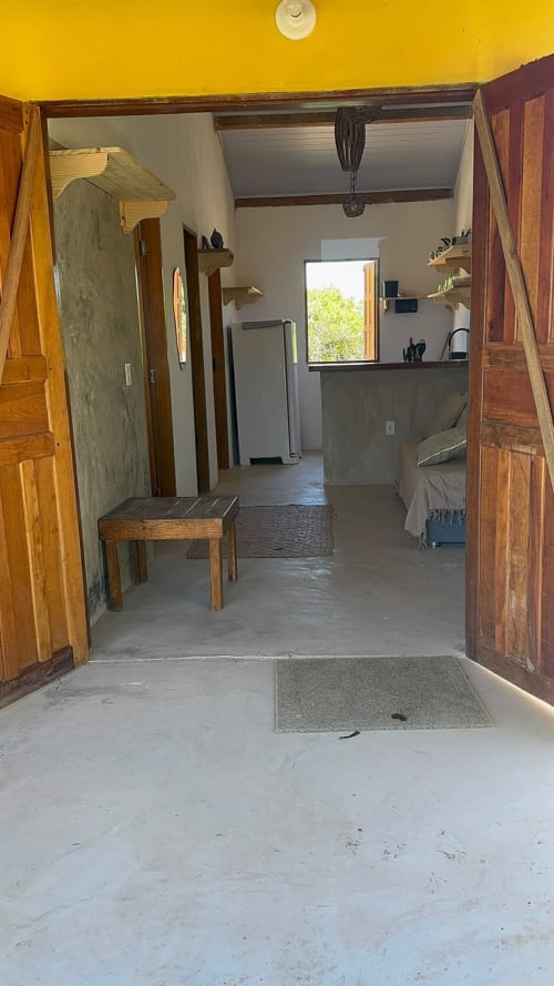 Entrada do apartamento Caraiva Cadu House com portas de madeira, teto amarelo, sala de estar mobiliada e piso de concreto
