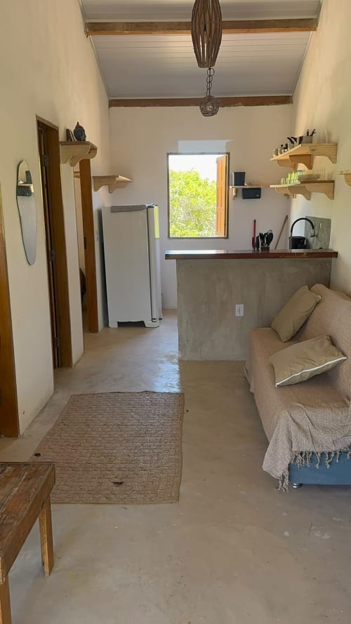 Interior do apartamento Caraiva Meynha com bancada de concreto, prateleiras de madeira, sofá bege e janela com vista para a vegetação