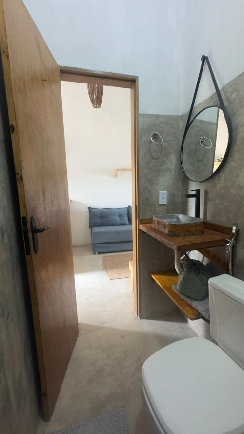 Banheiro moderno da Caraiva Cadu House com cuba de apoio, espelho redondo, vaso sanitário e vista da porta aberta para o quarto