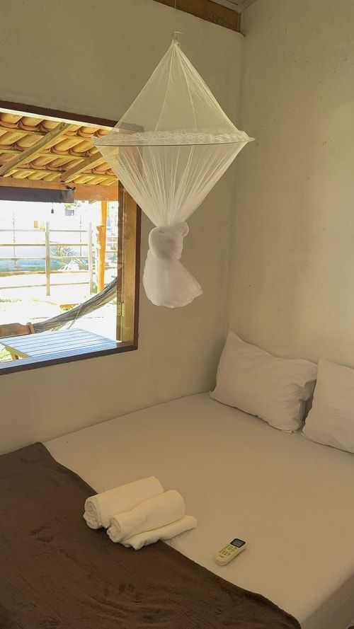 Quarto à beira-mar com dossel de mosquiteiro branco, almofadas, toalhas enroladas e vista para o oceano pela janela em Caraiva Cadu