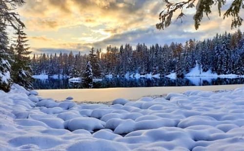 Paesaggio invernale nevoso con lago ghiacciato, foresta di conifere e cielo al tramonto dorato vicino a Villa Rosati a Sanzeno, Trentino-Alto Adige