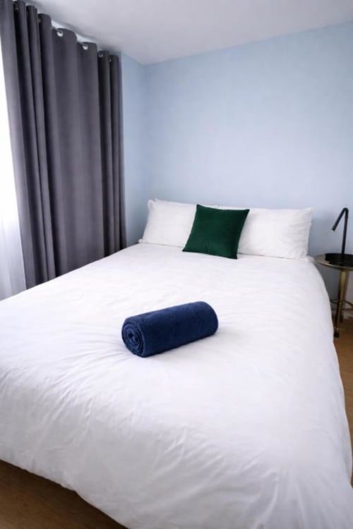 Bright bedroom at Victoria House Cozy Rooms, London B&B with white bed, grey curtains, green pillow, and blue towel