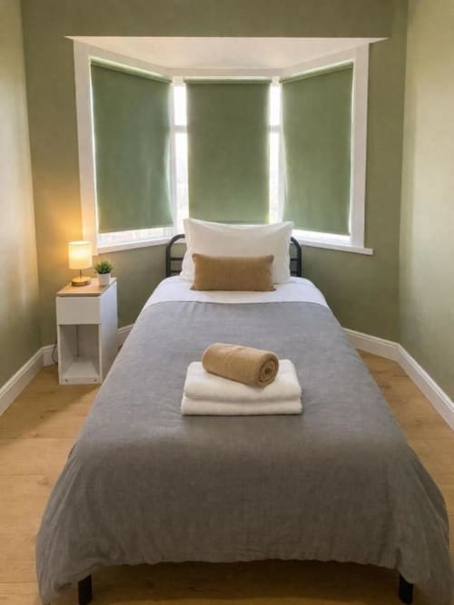Cozy bedroom at Victoria House, London B&B, featuring twin bed with grey linens, green roller blinds, white nightstand, and