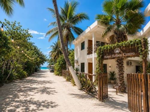 Entrada frente al mar del Hotel Holbox Centro con edificio blanco, palmeras y sendero arenoso hacia el Caribe turquesa