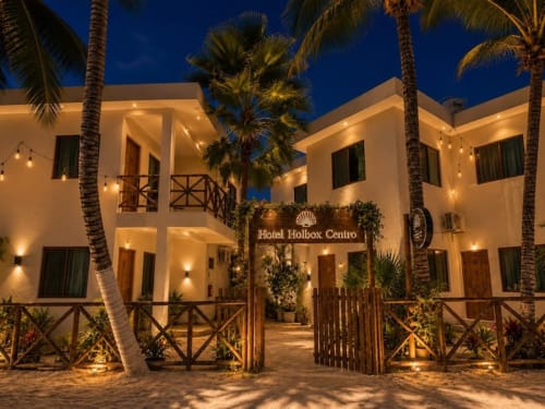 Fachada del Hotel Holbox Centro por la noche con edificios blancos iluminados, palmeras y barandales de madera en México