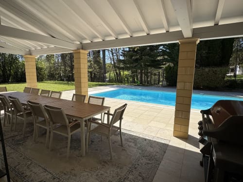 Überdachte Terrasse mit Esstisch mit Blick auf den blauen Swimmingpool und grüne Gärten des Domaine de Bize in Mirepoix, Ariège
