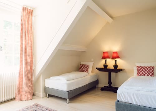 Helles Dachzimmer im Domaine de Bize Mirepoix mit zwei Betten, roten Gingham-Vorhängen und rotgetonten Lampen in Ariège, Frankreich