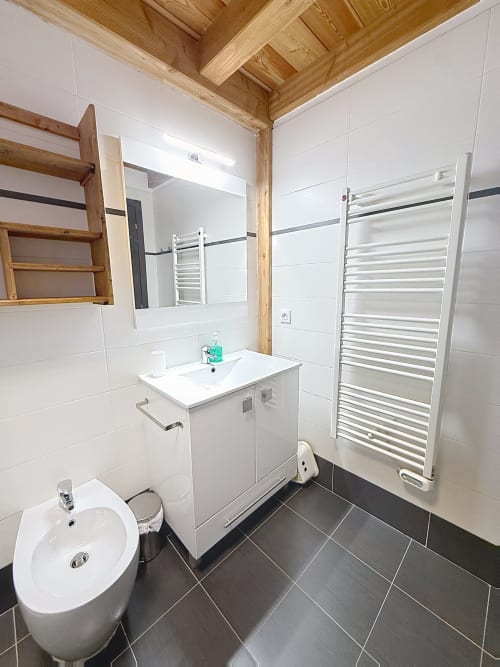 Salle de bain moderne avec vasque blanche, sèche-serviettes chauffant, carrelage foncé et plafond à poutres en bois aux Tanières de
