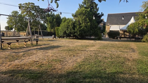Jardin rustique du Manoir de la Bégaudière avec balançelles, pelouse verdoyante et bâtiments en pierre sous ciel dégagé