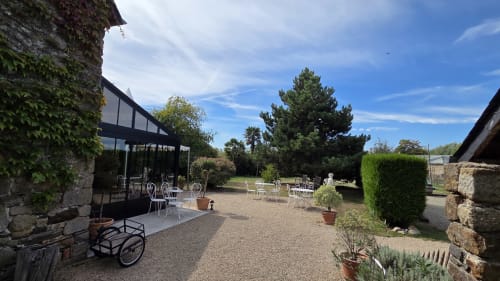Terrasse extérieure du Manoir de la Bégaudière avec chaises blanches, bâtiments en pierre et jardins luxuriants à Mont-Dol, France.
