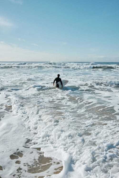 Surfeur en combinaison noire portant une planche blanche entrant dans les vagues à la plage El Ghazouane, Maroc près de l'hôtel Eucalyptus