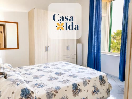 Camera luminosa a Casa Ida a Vietri con biancheria azzurra decorata, armadio crema e finestre con vista mare e tende blu.
