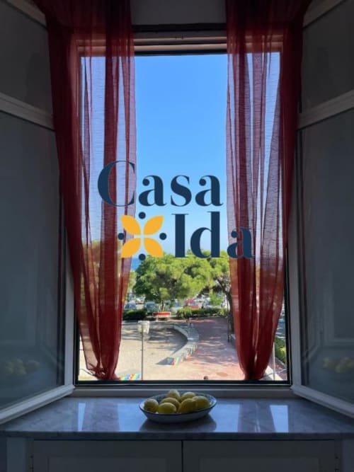Vista dalla finestra di Casa Ida con tende rosse sulla strada Vietri, fiori gialli in una ciotola sul davanzale