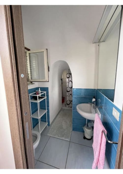 Bagno di Casa Ida Vietri con pareti rivestite di piastrelle blu, lavabo bianco, wc, specchio e mensola con asciugamano rosa