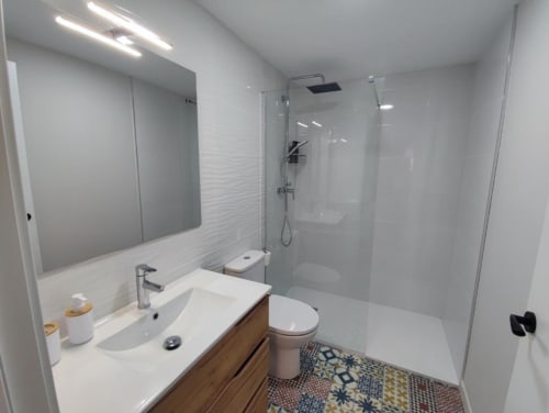 Baño moderno con lavabo blanco, inodoro, ducha con mampara de cristal y suelo con azulejos estampados en apartamento rural de Bronchales