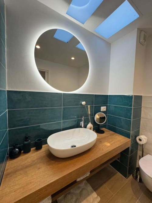 Bagno moderno con specchio circolare illuminato, lavabo bianco a bacinella, piastrelle turchesi, vanità in legno e lucernario a Casa Ida,
