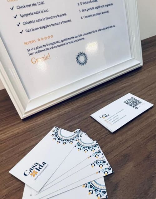 Materiali di branding Casa Ida e biglietti informativi su superficie in legno con codice QR e linee guida per gli ospiti in italiano.