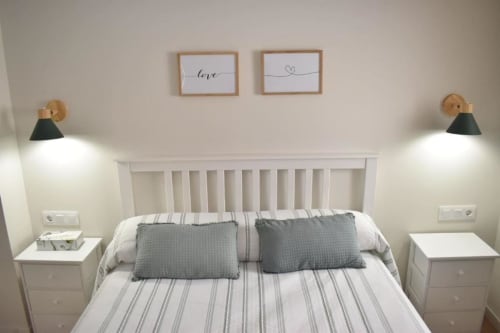 Dormitorio luminoso con cama de marco blanco, almohadas grises, ropa de cama rayada, apliques de pared y cuadros de amor en Casa El Abuelo