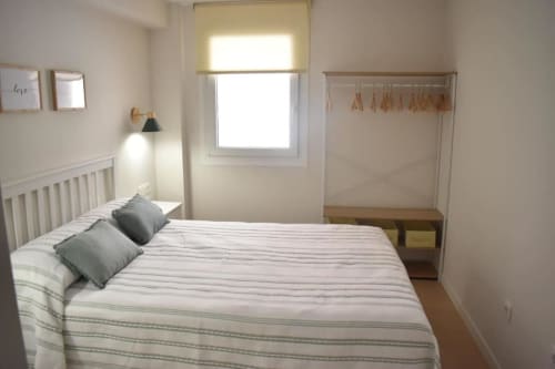 Dormitorio luminoso en casa rural española con ropa de cama a rayas, almohadas grises, ventana con persiana amarilla, lámpara de pared