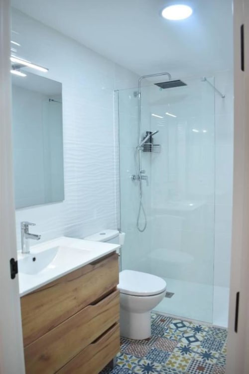 Baño moderno con tocador de madera, lavabo blanco, inodoro, ducha con mampara de vidrio y piso con azulejos estampados en Casa El Abuelo