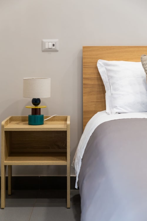 Modern bedroom at Darna Rooms Naples featuring wooden bed, nightstand with lamp, and light gray wall with outlet