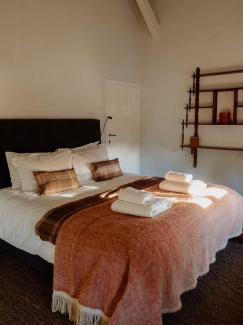 Cozy bedroom at Castanea Business & Suites with cream bedding, terracotta blanket, rolled towels, and wooden shelving in