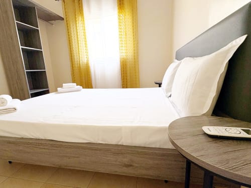 Chambre moderne chez DakarByDays avec draps blancs, tête de lit grise, meubles en bois, rideaux jaunes et étagères