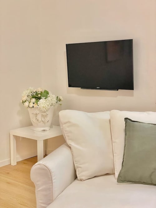 Soggiorno beige con poltrona bianca, cuscino verde, TV a parete e tavolino con fiori bianchi a Bologna