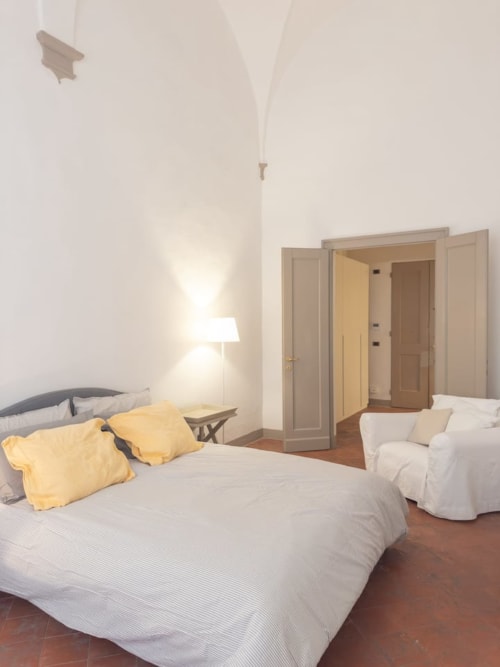 Camera luminosa nel palazzo storico di Bologna con letto matrimoniale, cuscini gialli, poltrona bianca, pavimenti in legno e porte aperte