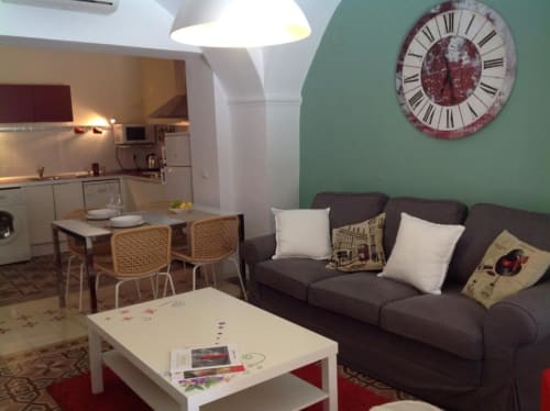 Sala de estar de apartamento moderno de 1 dormitorio en Cáceres con sofá oscuro, mesa de comedor y cocina, decorado con reloj de pared y