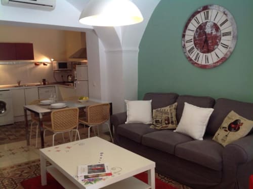 Moderno salón con sofá oscuro, reloj de pared, comedor y cocina en el apartamento Plaza de Santiago, Cáceres