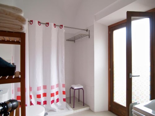 Baño moderno en apartamento Plaza de Santiago, Cáceres, con cortina de ducha a cuadros rojos, inodoro y puerta cristal a balcón