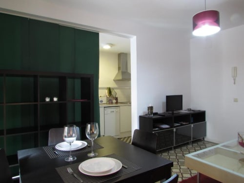 Apartamento moderno de planta abierta con área de comedor, sala de estar con TV, cocina visible y lámpara colgante púrpura