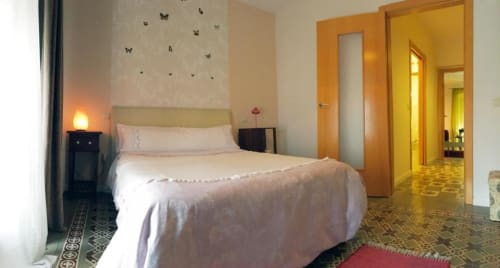 Luminoso dormitorio en apartamento de Cáceres con cama queen, cabecero en tonos salvia, arte mural de mariposas, iluminación cálida y puertas de madera
