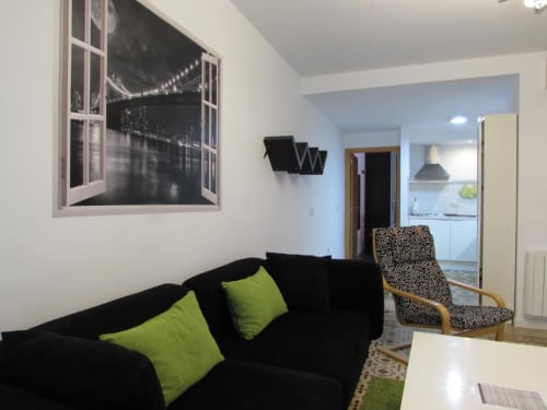 Sala de estar moderna en apartamento Plaza de Santiago, Cáceres: sofá negro con cojines lima, vista a cocina, arte urbano en ventana
