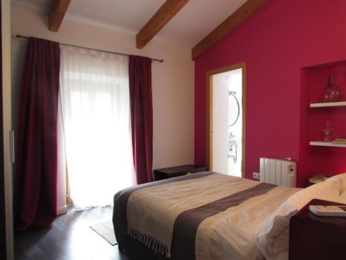 Dormitorio luminoso en apartamento de Cáceres con paredes magenta, vigas de madera, cama matrimonial y acceso a balcón