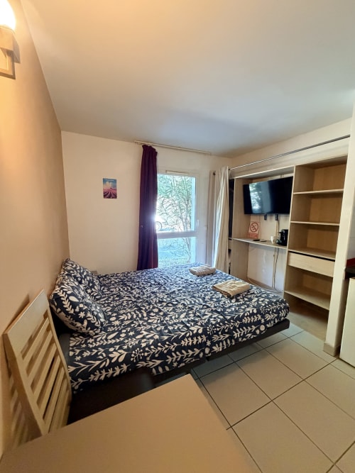 Le Clos d'Aix Studio 11 bedroom with patterned bed, garden view balcony, TV, kitchenette, and wooden shelving in Southern