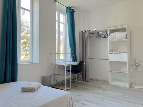 Chambre moderne à la Conciergerie-lilloise avec lit, bureau, penderie blanche, vue sur la ville et rideaux bleu canard
