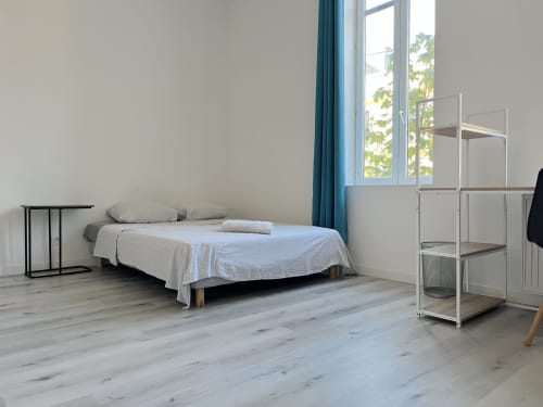 Chambre épurée à la Conciergerie-lilloise : lit, étagères, parquet, vue sur la ville à travers des rideaux bleu-vert