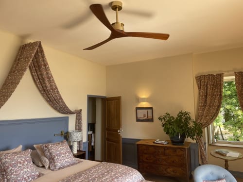 Elegant bedroom at Château de Vaux Morvan with blue headboard, patterned curtains, ceiling fan, wooden dresser, and garden