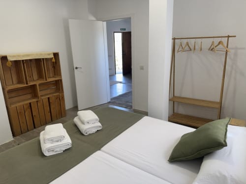 Espaïós dormitori de l'apartament a Séncies Can Girona, Sitges amb mobles de fusta, llençols blancs, coixí verd i peces de roba