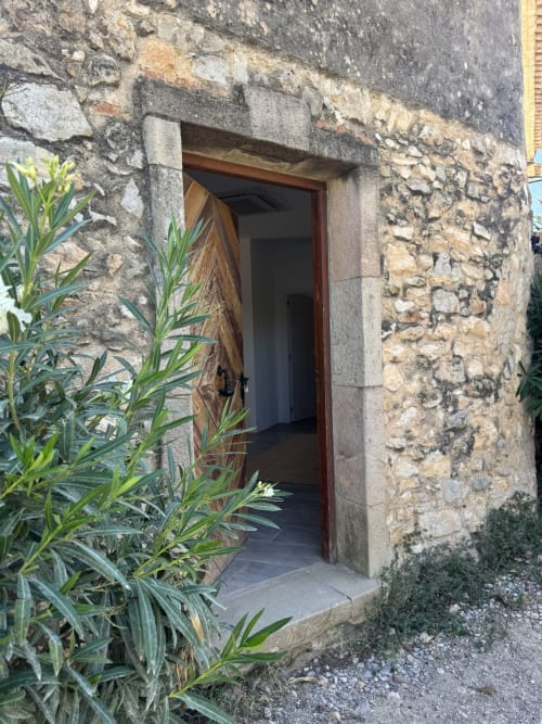 Entrada de pedra gastada a l'apartament Séncies Can Girona a Sitges, amb porta de fusta i plantes verdes
