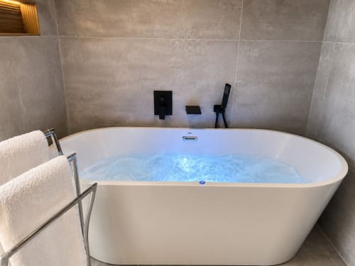 Bañera jacuzzi moderna con agua iluminada en azul en lujoso baño de apartamento en Baiona, con azulejos grises y toallas blancas