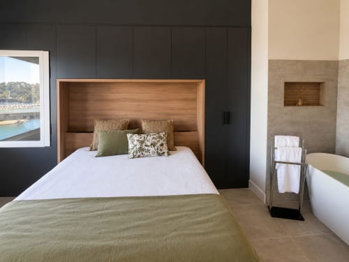 Dormitorio moderno con cabecera de madera, ventanal con vistas al mar y baño en suite en el hotel Holibai de Baiona