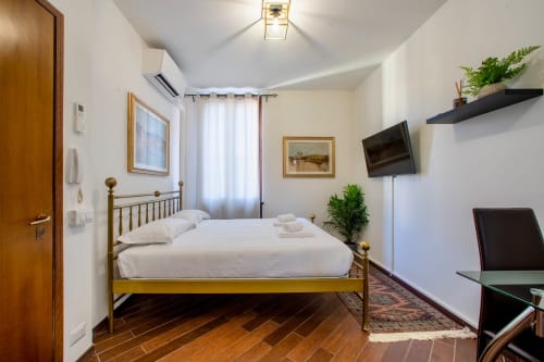 Camera da letto luminosa con letto in ottone, biancheria bianca, pavimento in legno, TV e piante in vaso nell'appartamento di Verona